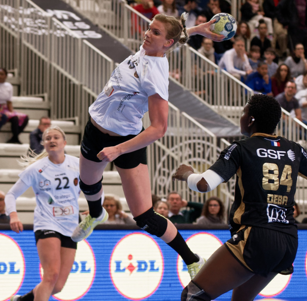Eine Gruppe von Frauen, die auf einem Platz Handball spielen, mit einem Ball in der Luft, Zuschauern auf den Stufen und einem "Handball-Weltmeisterschaft 2019 - Tag 2"-Schild im Hintergrund.