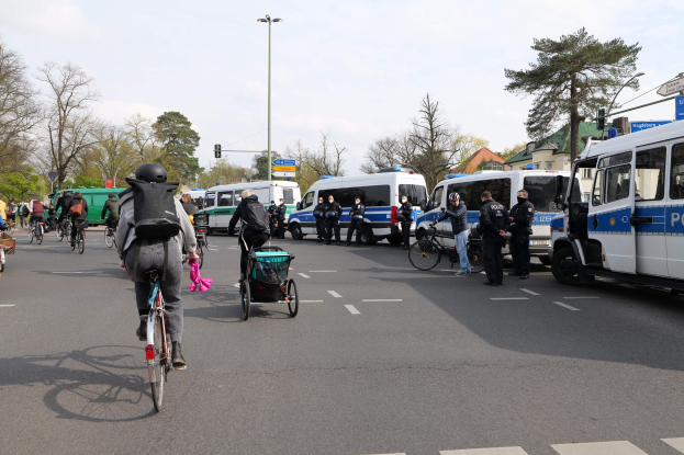 Eine Gruppe von Menschen, die auf Fahrrädern eine Straße entlangfahren, mit Polizeiwagen, anderen Fahrzeugen, Bäumen, Laternen, Verkehrsampeln, Schildern, Gebäuden und einem bewölkten Himmel.