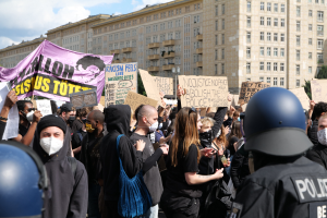 Gruppe maskierter Demonstranten mit Schildern vor einem Gebäude, mit zwei Polizisten in Helmen rechts daneben, vor einem Hintergrund aus Bäumen und bewölktem Himmel.