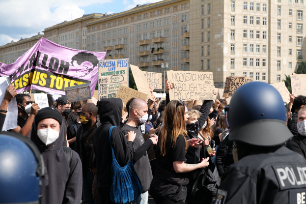 Gruppe maskierter Demonstranten mit Schildern vor einem Gebäude, mit zwei Polizisten in Helmen rechts daneben, vor einem Hintergrund aus Bäumen und bewölktem Himmel.