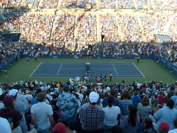 Eine große Menschenmenge in einem Stadion bei einem Tennismatch, mit Spielern auf dem Platz und Zuschauern in den Rängen, die eine elektrisierende Atmosphäre schaffen.