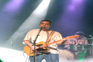 Ein Mann spielt eine E-Gitarre auf der Bühne, singt in ein Mikrofon, während ein Schlagzeuger hinter ihm spielt.