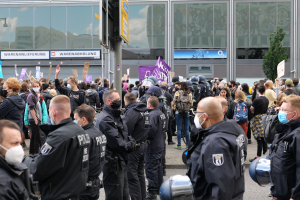 Eine große Gruppe von Menschen vor einem Gebäude stehend, einige mit Schildern und Helmen, mit einem Mast mit einer Schautafel im Vordergrund und einem Baum im Hintergrund, scheinbar protestierend.