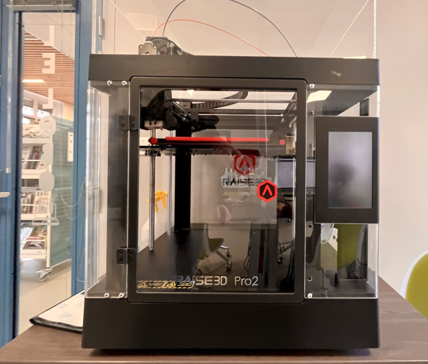 Ein 3D-Drucker auf einem Holztisch mit einer Glastür links und einem Stuhl rechts, vor Regalen mit verschiedenen Gegenständen im Hintergrund.