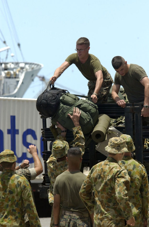 Männer in Militäruniformen laden Gepäck auf einen Lastwagen, mit einem Container, Schiff und einem klaren blauen Himmel im Hintergrund.