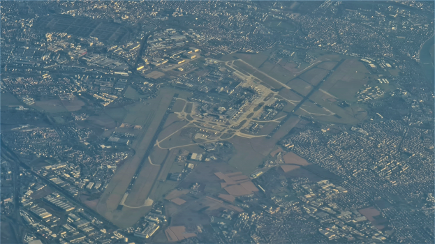 Luftaufnahme eines Flughafens umgeben von städtischer Infrastruktur, mit sichtbaren Gebäuden, Straßen und Text unten.