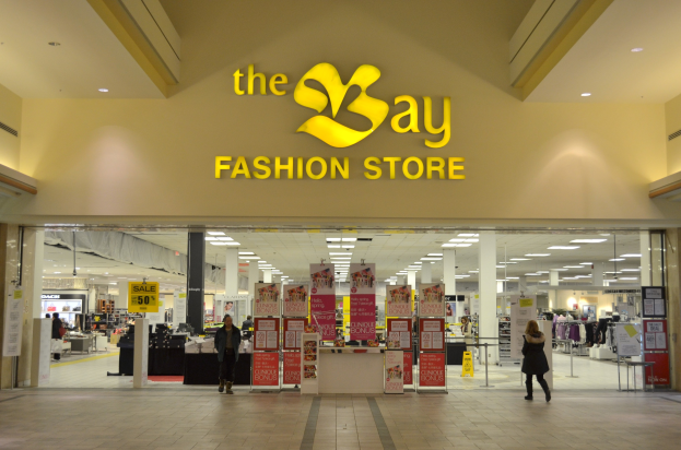 Außenansicht eines Bay Fashion Store in einem Einkaufszentrum mit Passanten, Wandtexttafeln, Säulen, Deckenleuchten und Hintergrundauslagen.
