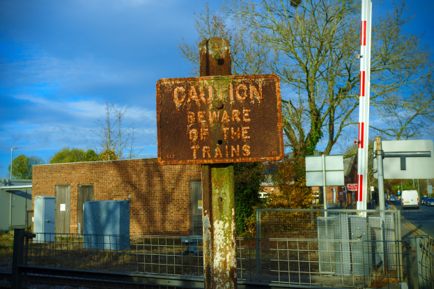 Vorsichtsschild auf einem Bahngleis umgeben von einem Metallzaun, Bäumen, Strommasten, einem Gebäude mit Fenstern, Containern, einem Straßenmast, Fahrzeugen auf einer Straße und einem bewölkten Himmel.