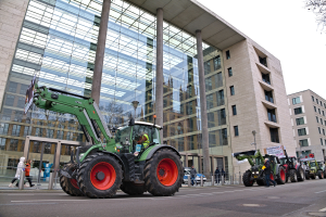 Eine Gruppe von Traktoren fährt auf einer Straße vor einem Gebäude, mit Menschen auf dem Gehweg und einem Baum auf der rechten Seite, während es scheint, dass sie an einer Demonstration teilnehmen.
