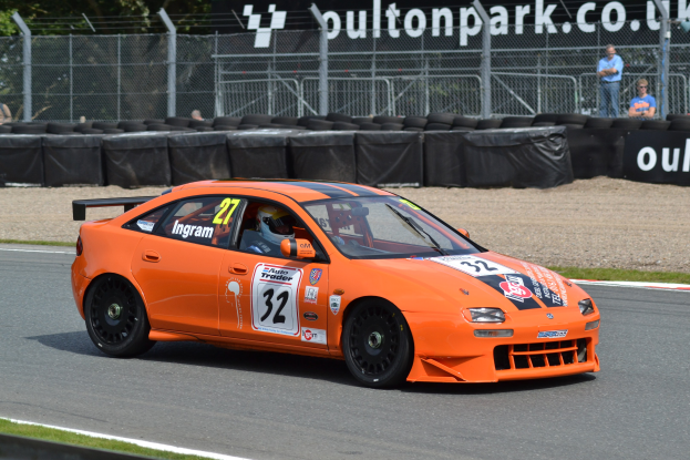 Ein oranges Rennauto auf einer Rennstrecke mit einem Helmträger am Steuer, umgeben von Reifen, Absperrungen, Zuschauern, Bäumen und einem Banner im Hintergrund.