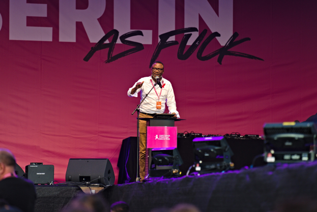 Ein Mann in einem wei├čem Hemd und braunen Hosen steht auf einer Bühne und h├Ąlt ein Mikrofon in der Hand, vor ihm eine rosafarbene Flagge mit der Aufschrift "Berlin as Fuck".