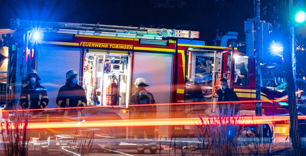 Eine Gruppe von Feuerwehrmännern in Helmen und Schutzausrüstung steht vor einem beleuchteten Feuerwehrauto bei Nacht, mit Pflanzen im Vordergrund, einem Verkehrszeichen auf der rechten Seite und Bäumen im Hintergrund unter einem dunklen Himmel.