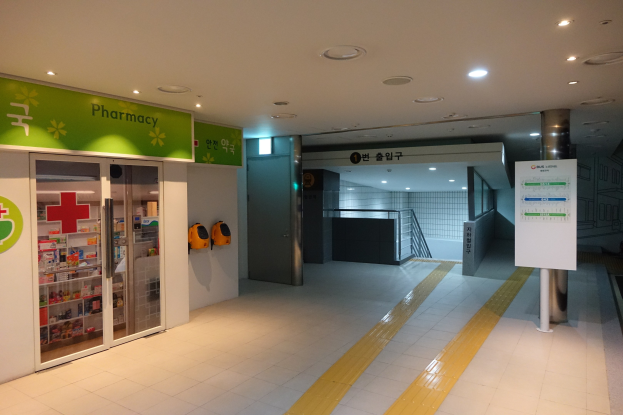 Eingang einer Apotheke mit einem "Apotheke"-Schild links, einer Tafel mit Text rechts und beleuchteten Lampen über dem Dach.