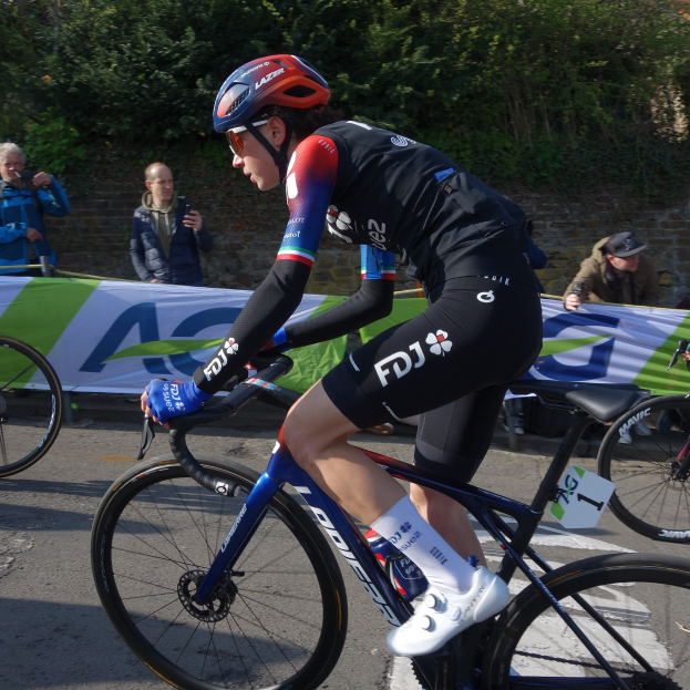 Eine professionelle Radrennfahrerin, Katie Archibald, fährt in einem Helm, einer Brille, Handschuhen und Schuhen auf einem Fahrrad eine Straße entlang neben einer Menschenmenge, mit einem Banner, einer Wand, Bäumen und einem Gebäude im Hintergrund.