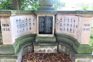 Ein Holocaust-Denkmal-Monument in einem jüdischen Friedhof in Berlin, das eine Tafel mit Text und Zahlen zeigt, umgeben von Bäumen, einem Zaun und verstreuten trockenen Blättern.