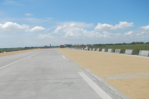 Eine Straße mit Betonbarrieren auf beiden Seiten, Fahrzeuge, die darauf fahren, und Bäume und Wolken im Hintergrund.