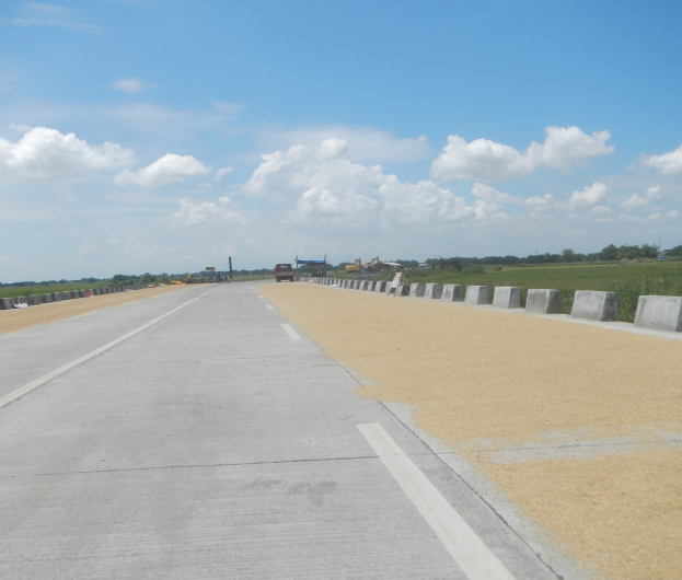 Eine Straße mit Betonbarrieren auf beiden Seiten, Fahrzeuge, die darauf fahren, und Bäume und Wolken im Hintergrund.
