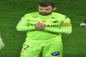 Ein Mann in einem gelben Torwarttrikot, der ein grünes T-Shirt und Shorts trägt, steht auf einem Fußballfeld und klatscht in die Hände, wobei ein weiteres Bein auf der linken Seite des Bildes zu sehen ist.