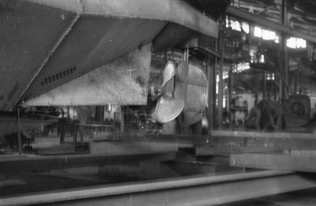 Ein Schwarz-Weiß-Foto eines Boots in der Fertigung in einer Fabrik, umgeben von Metallobjekten, Pfählen und Lichtern.