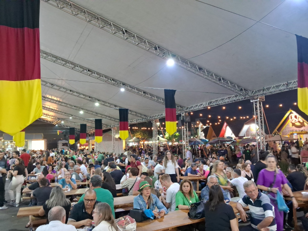 Eine große Gruppe von Menschen sitzt an Tischen unter einem Zelt beim Oktoberfest in München, Deutschland, einige stehen und Taschen sind auf den Tischen, sowie Zelte, Lichter und Fahnen im Hintergrund.