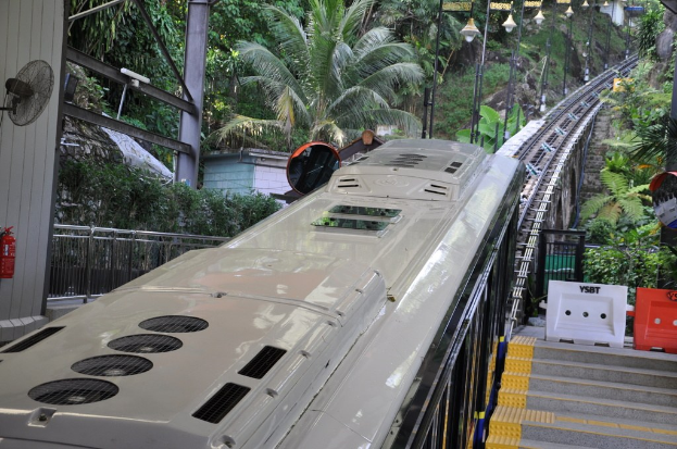 Ein Zug fährt auf einem Bahngleis mit Gebäuden, Strommasten, einem Ventilator, einem Feuerlöscher und einem Geländer auf der linken Seite und Objekten mit einer Treppe auf der rechten Seite, vor einem Hintergrund aus Bäumen.