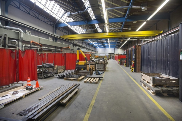 Großes Lagerinnere mit Holzpaletten, Metallobjekten, Verkehrskegeln, Maschinenfahrzeug, mit roten Vorhängen, Deckenrohren, Lampen und einer Vorhangwand auf der rechten Seite.