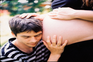 Ein Mann und eine Frau stehen nebeneinander, wobei der Mann zärtlich den Bauch der Frau berührt und auf ihr ungeborenes Kind schaut.