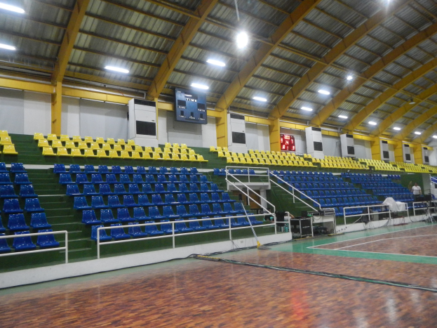 Eine geräumige Indoor-Sporthalle mit einem Holzboden, blauen und gelben Stühlen, Geländern, Treppen zum Sitzbereich, Bildschirmen an den Wänden, Deckenleuchten und einigen anwesenden Personen.