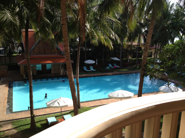 Großes Schwimmbad mit Menschen, umgeben von üppiger Vegetation, Schirmen, Stühlen, Häusern und einem klaren blauen Himmel, von einem Hotelbalkon aus betrachtet.
