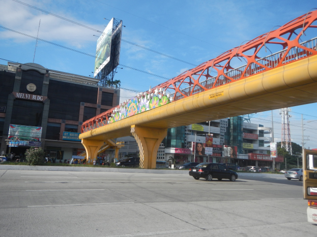 Eine Brücke spannt eine vielbefahrene Straße mit Autos, flankiert von Gebäuden, Werbetafeln, Strommasten, Kabeln und Bäumen unter einem bewölkten Himmel.