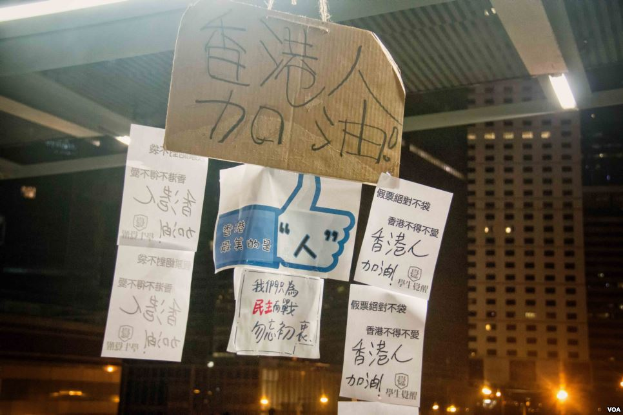 Eine Gruppe von Menschen steht vor einem Gebäude in der Nacht, beleuchtet von Stadtlichtern, mit mehreren Schildern von der Decke hängend, wahrscheinlich protestierend gegen eine Regierungsentscheidung, soziale Medien zu verbieten.