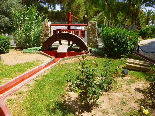 Minigolfplatz in einem Park mit Grünfläche, einer Straße mit Fußgängern und Schildern sowie einem klaren blauen Himmel.