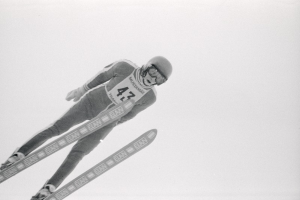 Ein Skispringer in voller Montur springt mitten in der Luft auf Skiern gegen einen klaren blauen Himmel.