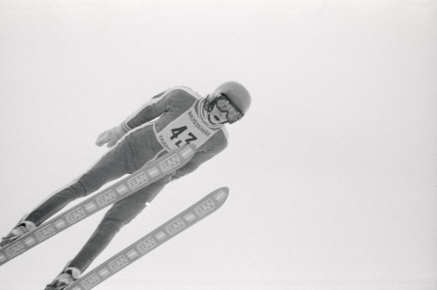 Ein Skispringer in voller Montur springt mitten in der Luft auf Skiern gegen einen klaren blauen Himmel.