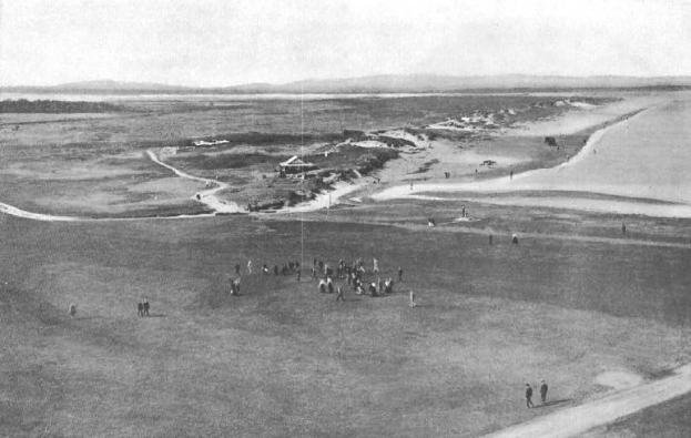 Schwarz-weiß-Foto von Golfern auf dem 18. Loch auf dem Royal Birkham Golf Club mit saftig grünem Rasen, verstreuten Häusern, sanften Hügeln und einem hellblauen Himmel im Hintergrund.