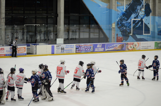 Gruppe junger Menschen, die Eis hockey auf einer Halle spielen, mit Helmen und Stöcken, sowie einem Zuschauer links und Bannern, Säulen und Glaswänden im Hintergrund.