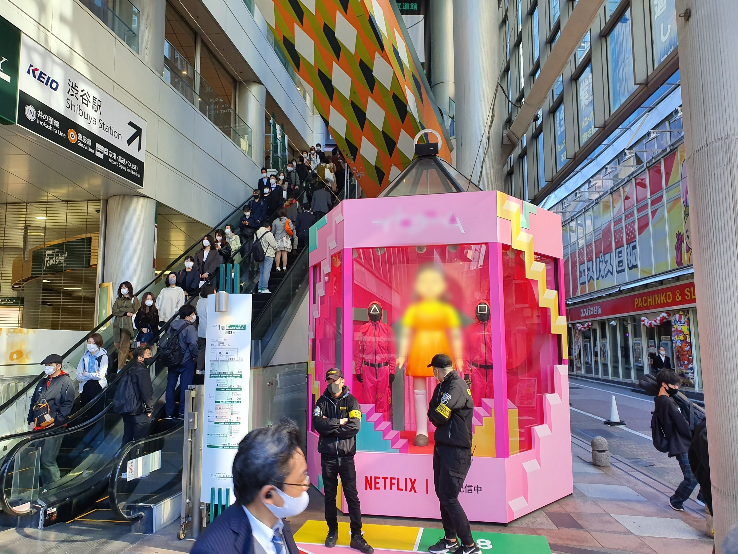 Eine Gruppe von Menschen steht vor einem Gebäude mit Glaswänden und Texttafeln, einige tragen Masken, während eine Rolltreppe mit Menschen zu sehen ist; ein pinker Kasten in der Mitte zeigt Schaufensterpuppen und trägt die Aufschrift "Netflix's new Netflix-Booth in Tokyo."