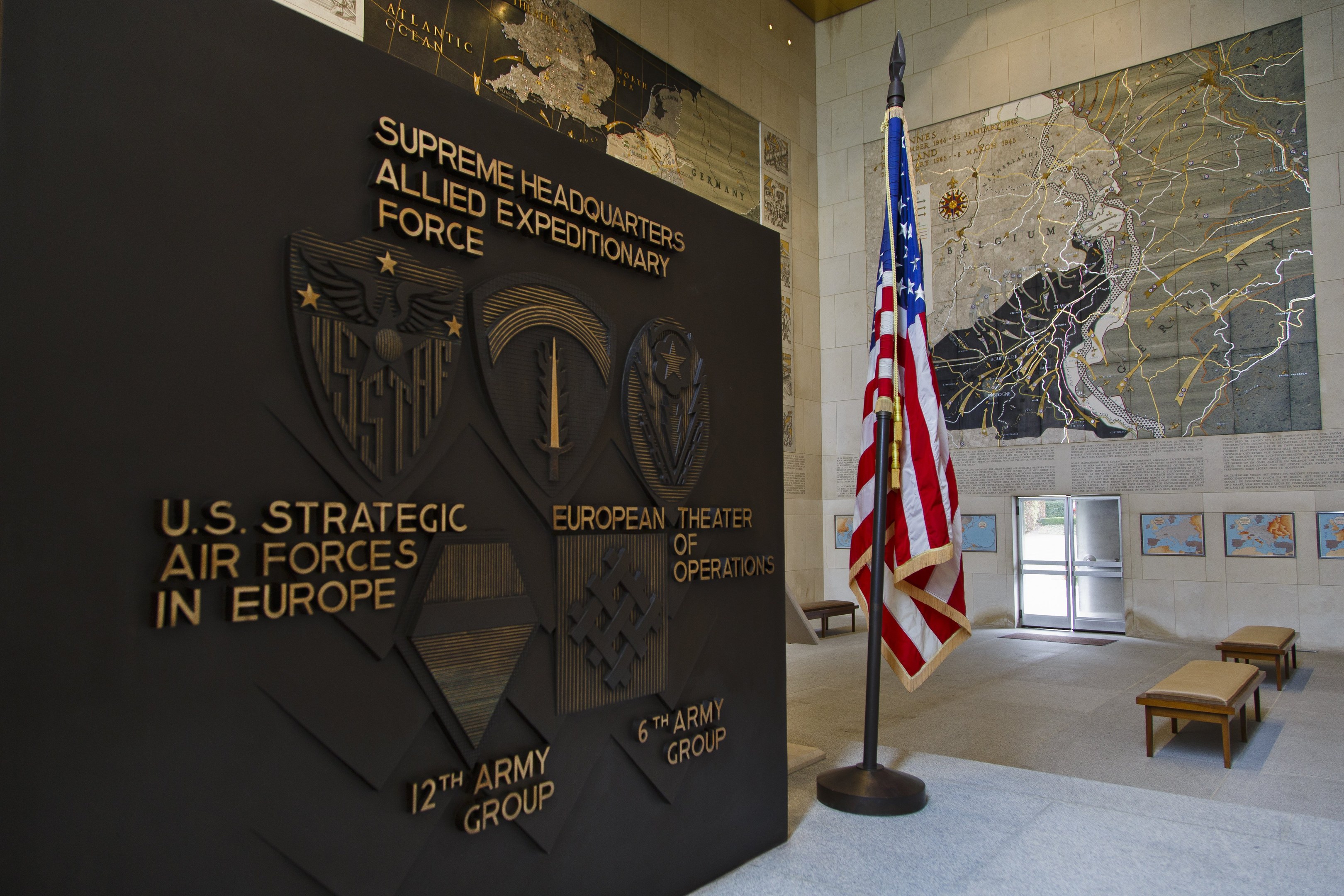 Eingang zum Hauptquartier der US-Strategischen Luftstreitkräfte in Europa mit einer amerikanischen Flagge, Bänken, einer Wandtafel, Fotorahmen und einer beleuchteten Decke, die zu einer Tür im Hintergrund führt.