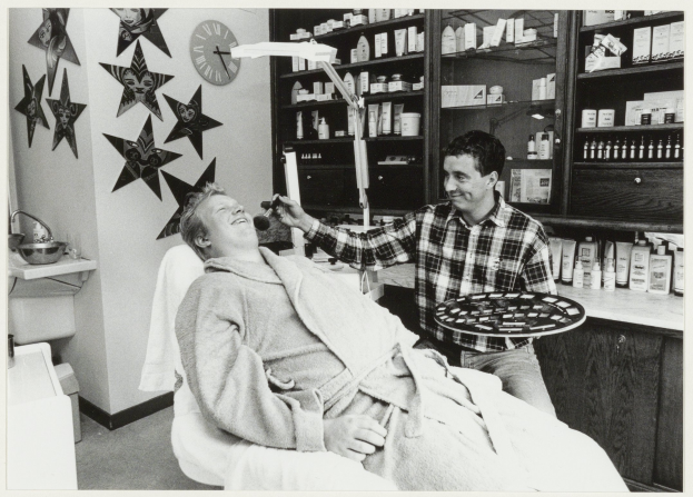 Ein Mann lächelt in einem Zahnarztstuhl, während ein anderer Mann neben ihm steht und ein Tablett hält, mit einem Waschbecken, Sternen, einer Uhr und Regalen mit Flaschen im Hintergrund.