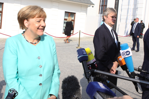 Bundeskanzlerin Angela Merkel spricht vor einem Gebäude mit Fenstern mit einer Gruppe von Menschen, einige halten Mikrofonstative, unter einem bewölkten Himmel.