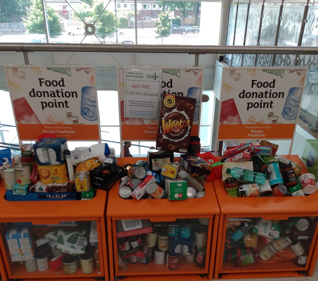 Orange Schübe mit Lebensmittelspenden, darunter Flaschen, Dosen und Kisten, mit Informationsplakaten, durch Glasfenster betrachtet, die Außenfahrzeuge, Gebäude, Bäume und Himmel zeigen.