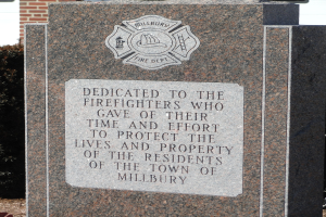 Denkmalstein mit einer Gedenktafel für Feuerwehrleute in Millbury, vor einem Backsteingebäude mit umliegenden Pflanzen und Bäumen.