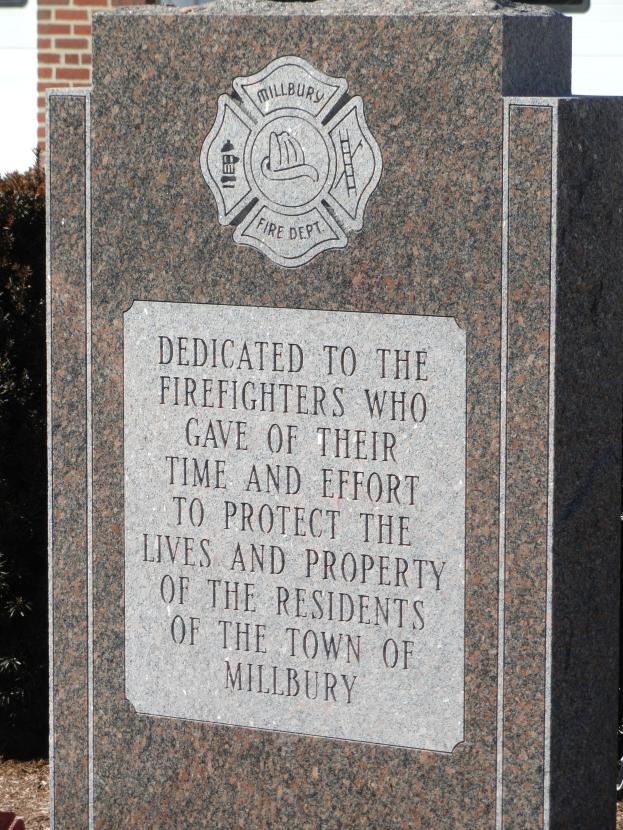 Denkmalstein mit einer Gedenktafel für Feuerwehrleute in Millbury, vor einem Backsteingebäude mit umliegenden Pflanzen und Bäumen.