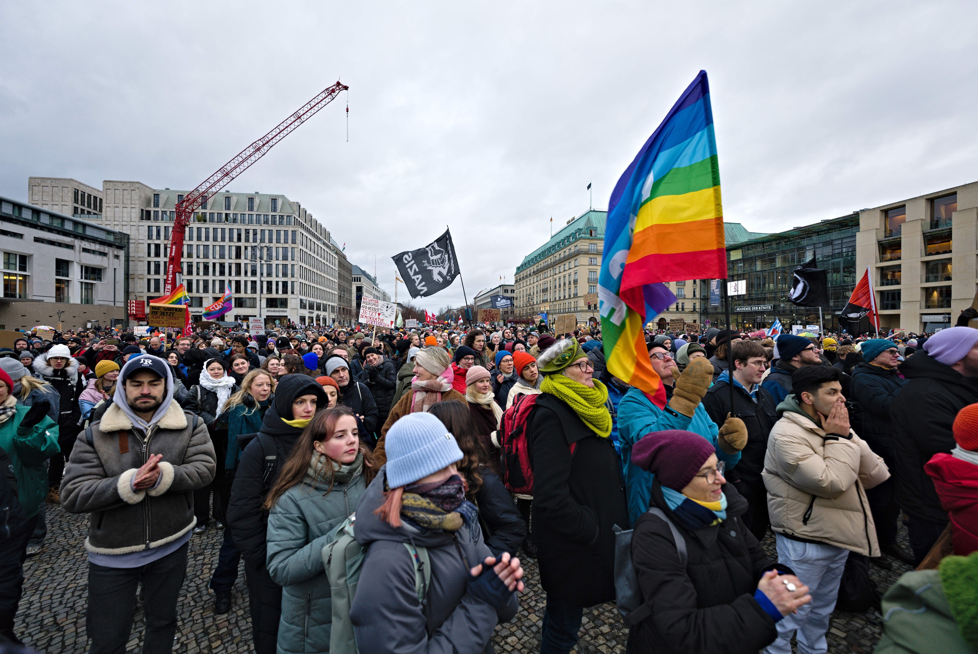 Eine große Gruppe von Menschen steht vor einem Gebäude und hält Fahnen und Schilder mit der Aufschrift "Lgbtq+ Rechte Demonstration in Berlin", einige tragen Mützen und Taschen, im Hintergrund sind Gebäude, ein Kran und ein bewölkter Himmel zu sehen.