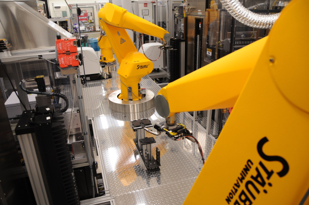 Gelber industrieller Roboter, der Maschine in einer Fabrikumgebung bedient, mit sichtbaren Rohren, Equipment und Deckenbeleuchtung.