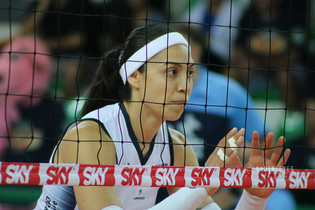 Eine weibliche Volleyballspielerin in Uniform ist im Einsatz auf dem Feld, mit einem Netz im Vordergrund und einem unscharfen Hintergrund, die eine entschlossene Miene zeigt.
