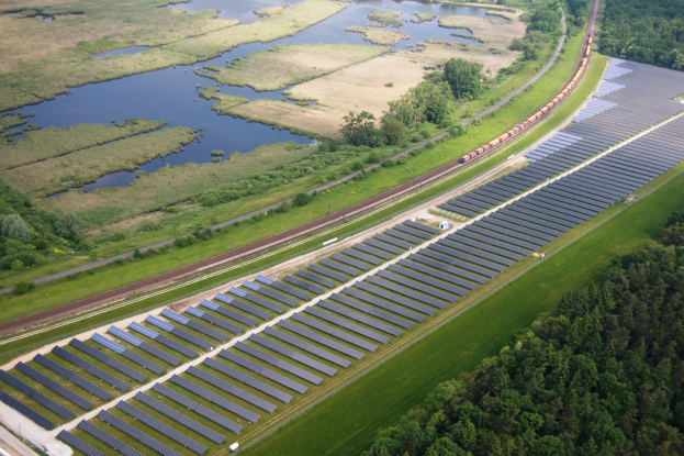 Luftaufnahme eines Solarparks mit Panelen, einem Zug auf einem Gleis, umgeben von Bäumen, Gras, Pflanzen und Wasser.