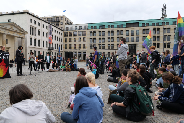 Eine Gruppe von Menschen, die auf dem Boden vor einer Menge sitzt, die Fahnen und Transparente hält, während einer Anti-Schwulen-Demonstration in Berlin. Im Hintergrund sind eine Statue, ein Mikrofon und Gebäude zu sehen.