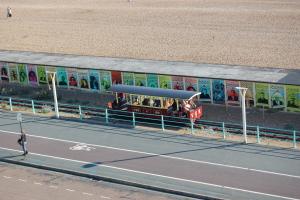 Ein kleiner Zug mit Passagieren fährt auf Schienen neben einem Strand, mit einer Person, die auf einem Fußweg links geht und einer Wand mit Plakaten im Hintergrund.
