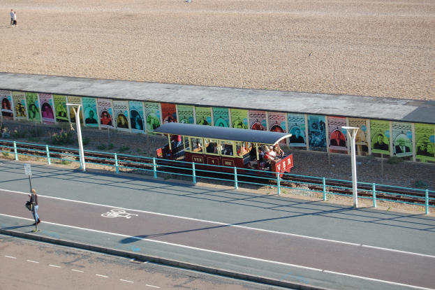 Ein kleiner Zug mit Passagieren fährt auf Schienen neben einem Strand, mit einer Person, die auf einem Fußweg links geht und einer Wand mit Plakaten im Hintergrund.